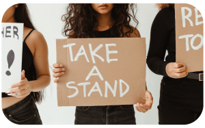 A woman with brown skin and long curly hair whose face is cropped holding a cardboard sign with the text "take a stand" surrounded by two other people cropped on either side in front of a white background
