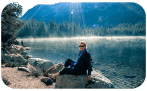 Photo of Claire, a white woman in navy wearing sunglasses and her blond hair up, sitting on a rock by a river with a mountain and trees in the background