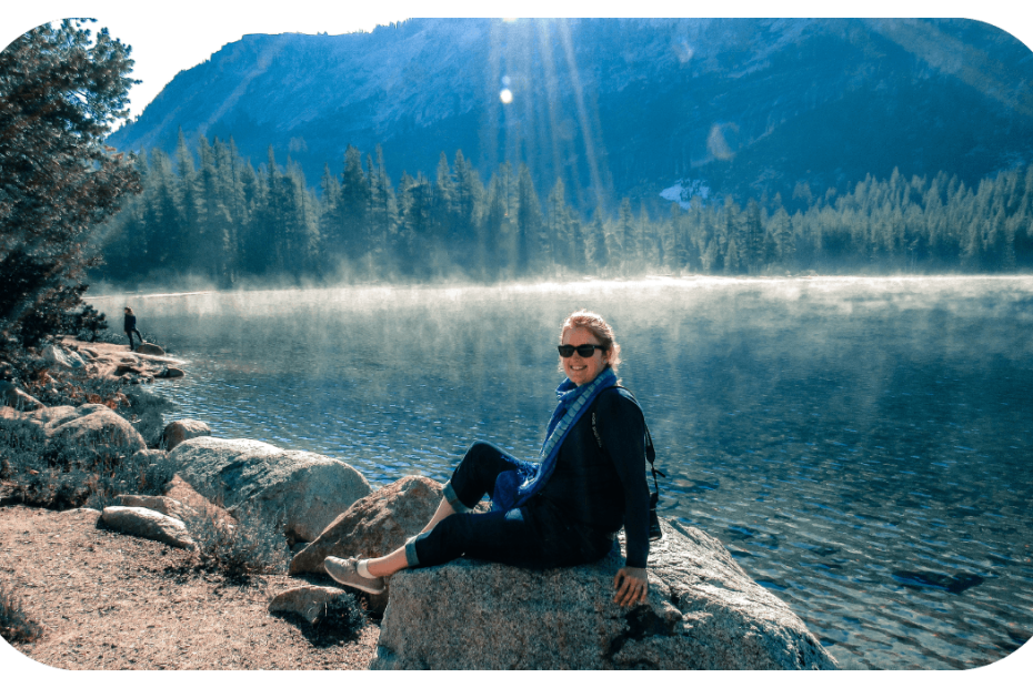 Photo of Claire, a white woman in navy wearing sunglasses and her blond hair up, sitting on a rock by a river with a mountain and trees in the background